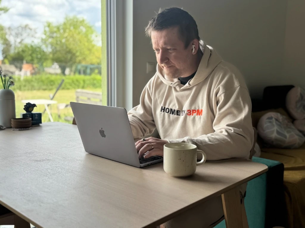 Michal in a Home by 3pm hoodie, working on a laptop at the kitchen table, garden window behind him